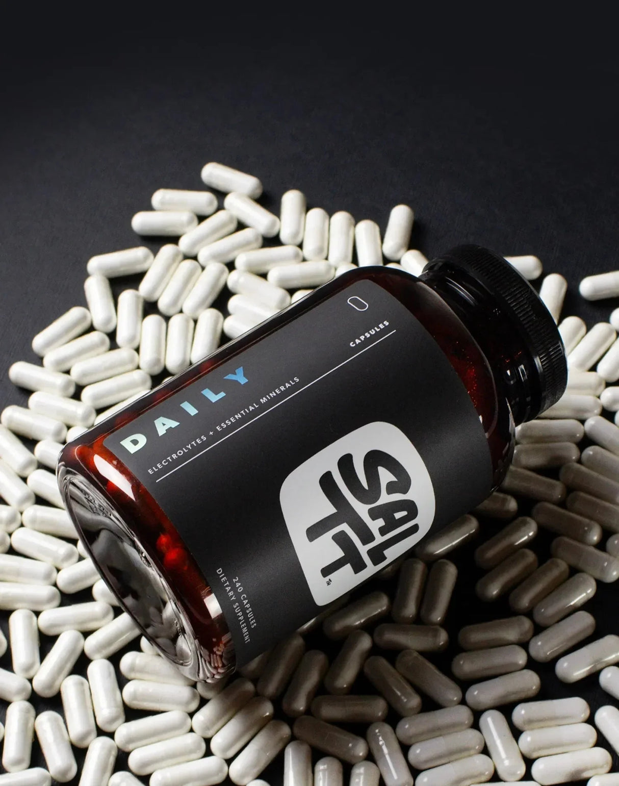 Dark amber bottle of Daily - Electrolytes + Minerals - 240 Caps by SALTT surrounded by white capsules on black surface.