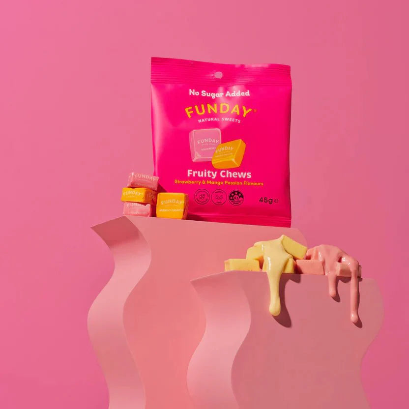 A pink packet of Funday Fruity Chews - 45g sits on a wavy pink surface with strawberry and mango chews stacked in front. Some chews are melting and dripping on the right side, all set against a solid pink background.