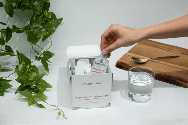Hand holding a pouch of CYMBIOTIKA Magnesium L-Threonate - 30 Pouches supplement next to a glass of water and green plant.