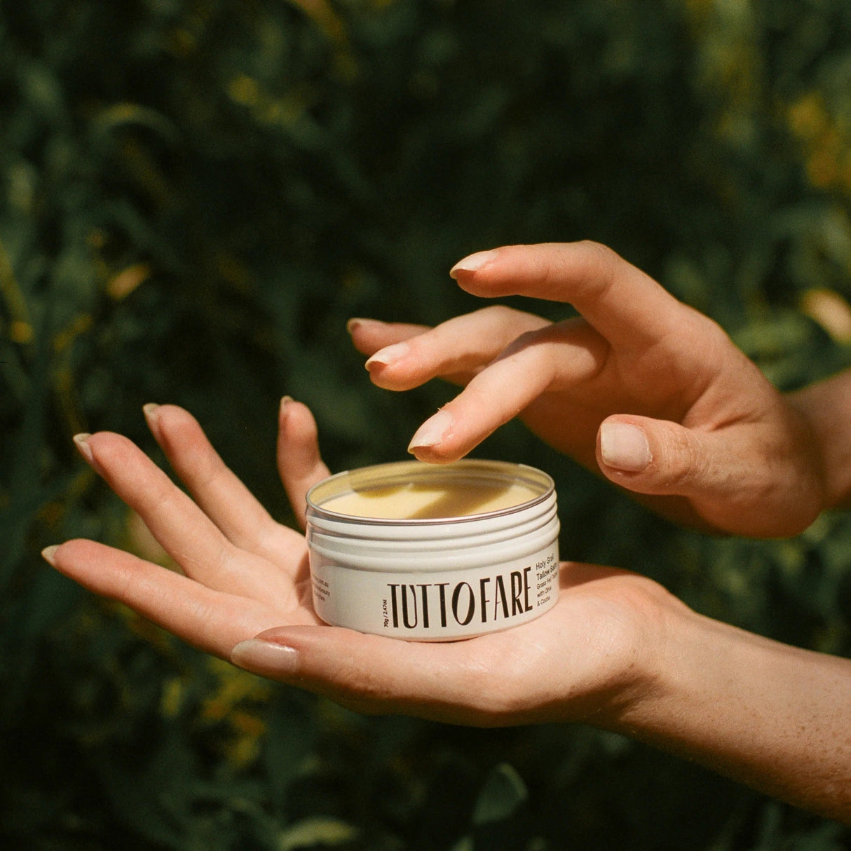 Hands holding a jar of Holy Grail Tallow Balm - 70g by Tuttofare with natural greenery background