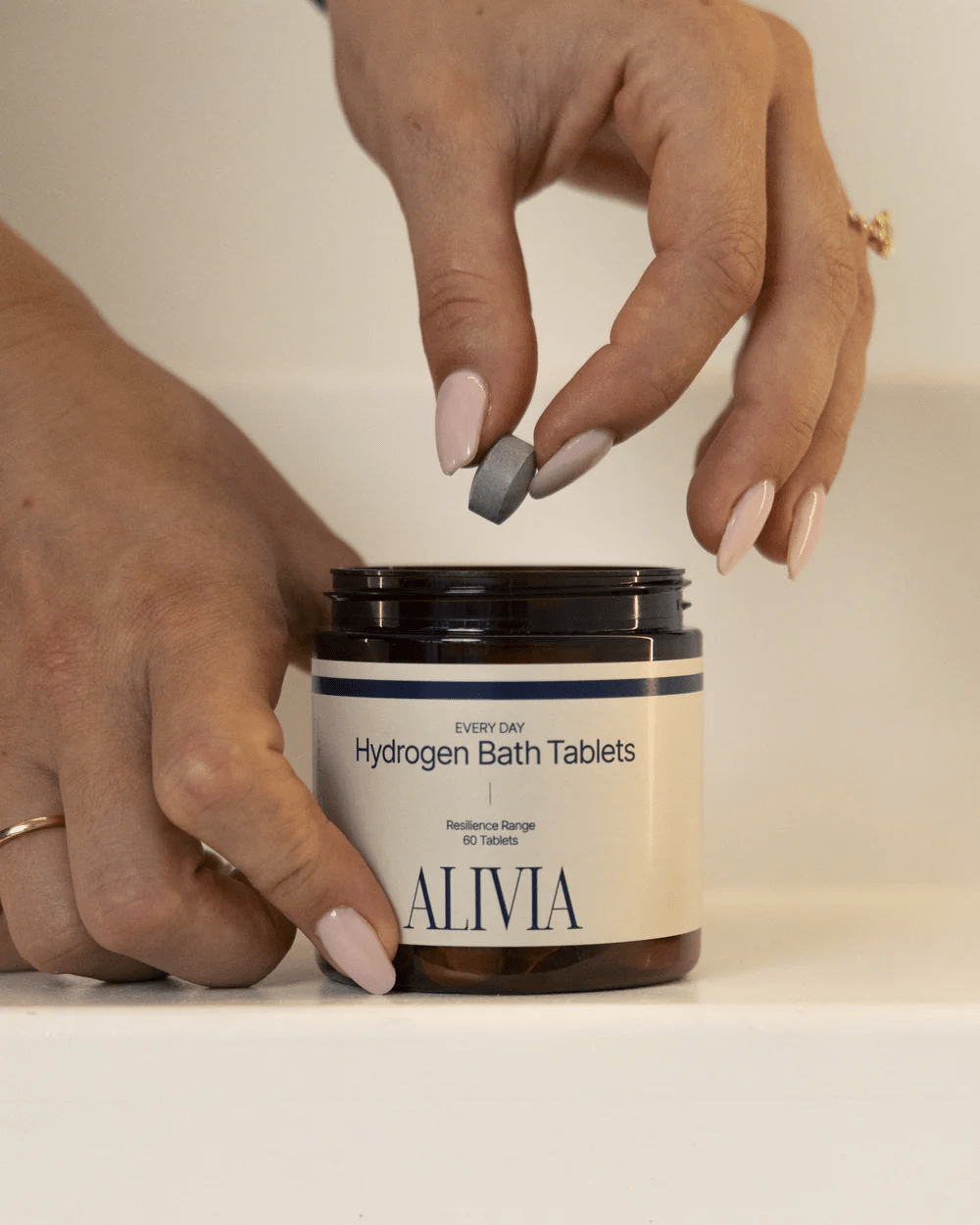 Hands holding a tablet above an open jar of Alivia Hydrogen Bath Tablets - 60pk from the New Arrivals collection.