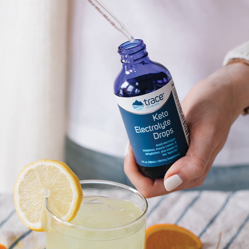 Hand holding blue bottle of Keto Electrolyte Drops -118ml with dropper above lemon water on striped cloth.