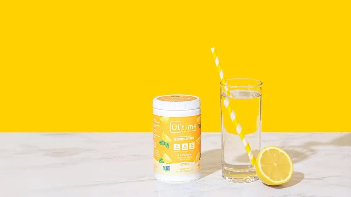 Lemonade Electrolyte Drink Mix - 90 Serves by Ultima Replenisher with glass of water and lemon on yellow background