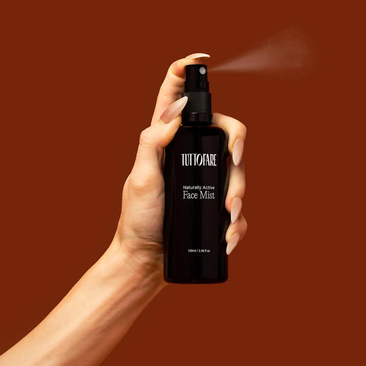 Hand holding Tuttofare Naturally Active Face Mist - 100ml bottle spraying fine mist on warm brown background