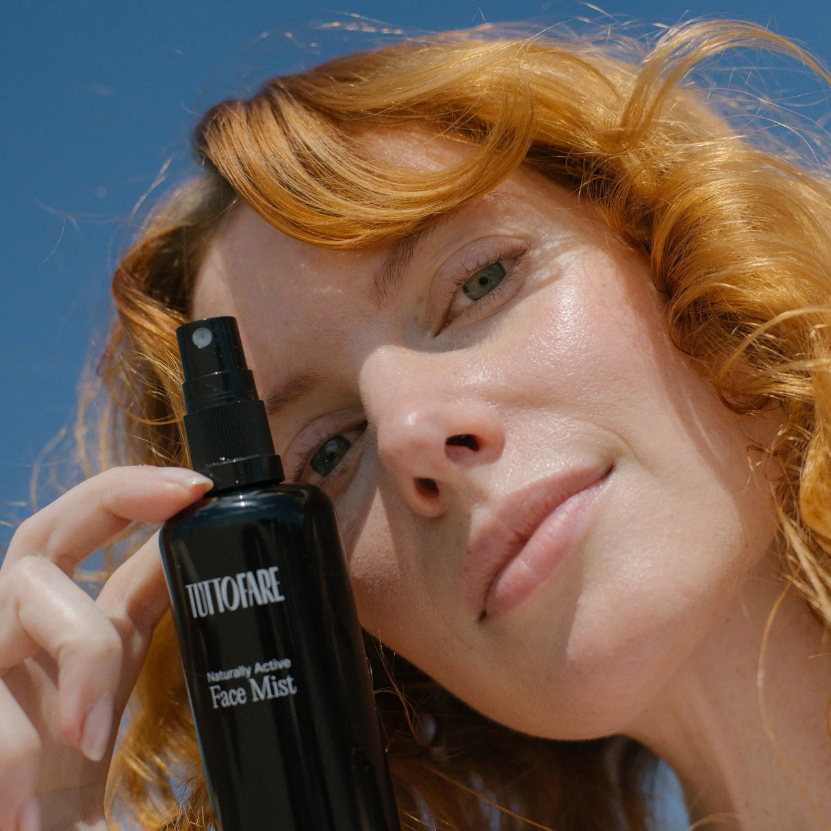 Woman with red hair holding a black bottle of Naturally Active Face Mist - 100ml by Tuttofare against a blue sky background