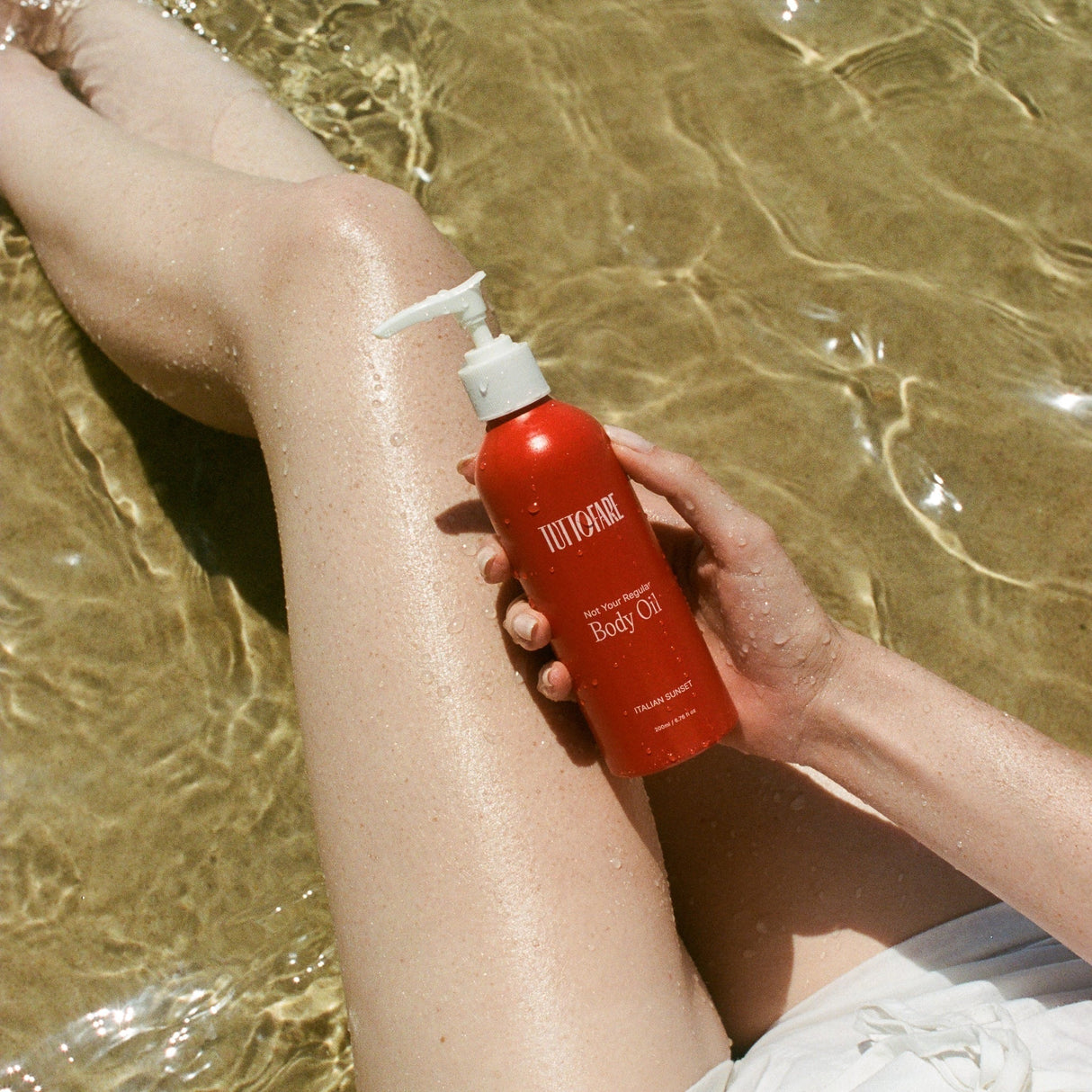 Hand holding Tuttofare Not Your Regular Body Oil - 200ml bottle near wet legs submerged in clear water outdoors