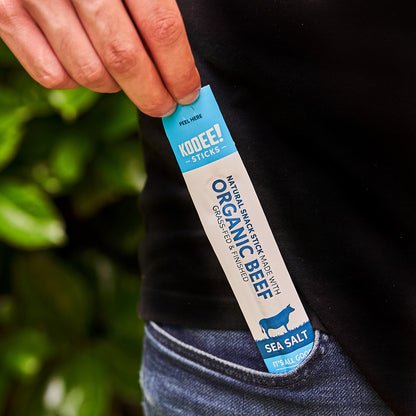 Hand holding a KOOEE! Organic Beef Stick Sea Salt snack from the Organic Beef Stick - Sea Salt - Box of 20 pack.