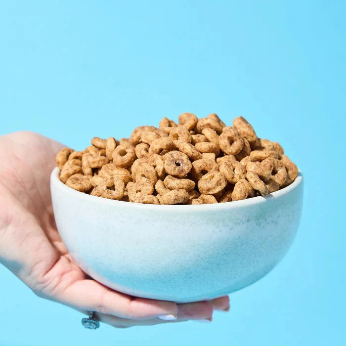 Hand holding a white bowl filled with Protein Cereal - Cinnamon Flavour - 250g by Low Carb Emporium against a blue background