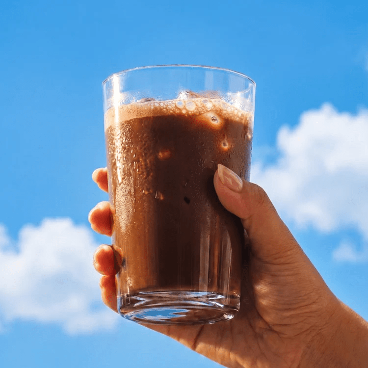 Hand holding a glass of frothy chocolate drink against a bright blue sky, showcasing Sodii’s Salty Chocolate - 30 Serve Tub for keto hydration.