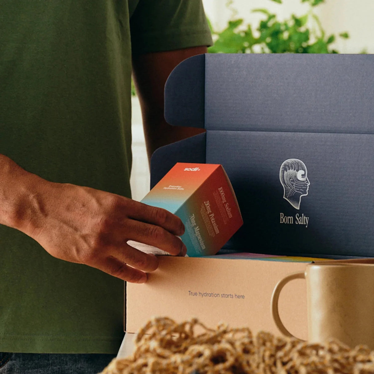 Person placing Sodii Salty Grapefruit - 30 Sachets box into Born Salty packaging with mug and woven mat nearby