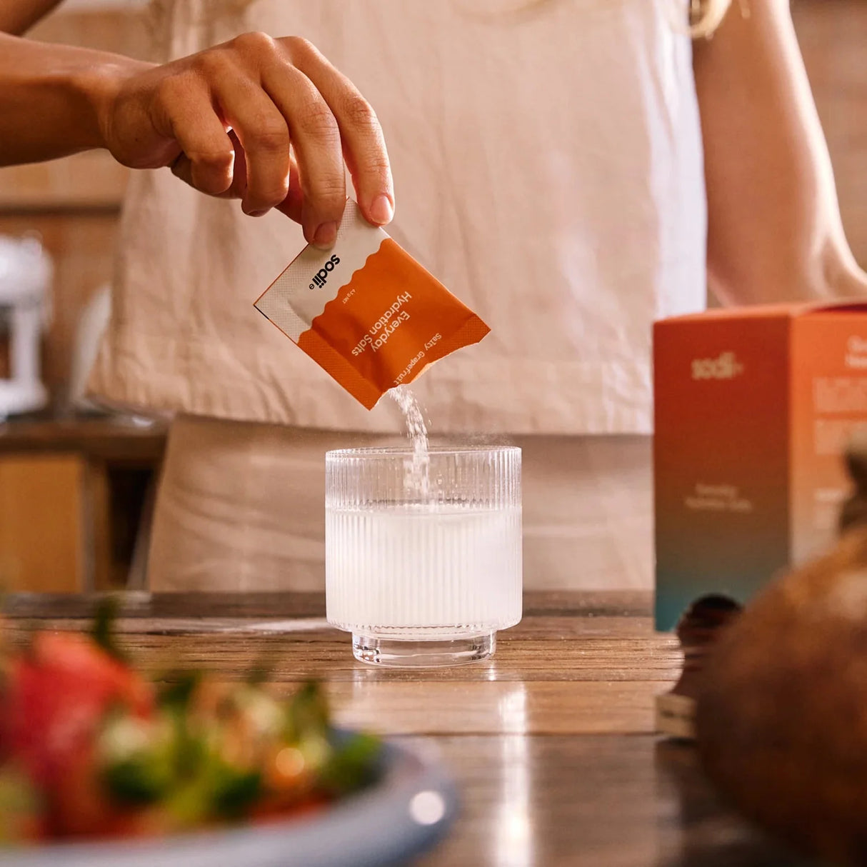 Person pouring Salty Grapefruit - 30 Sachets by Sodii electrolyte powder into a glass on a kitchen counter with fruit nearby