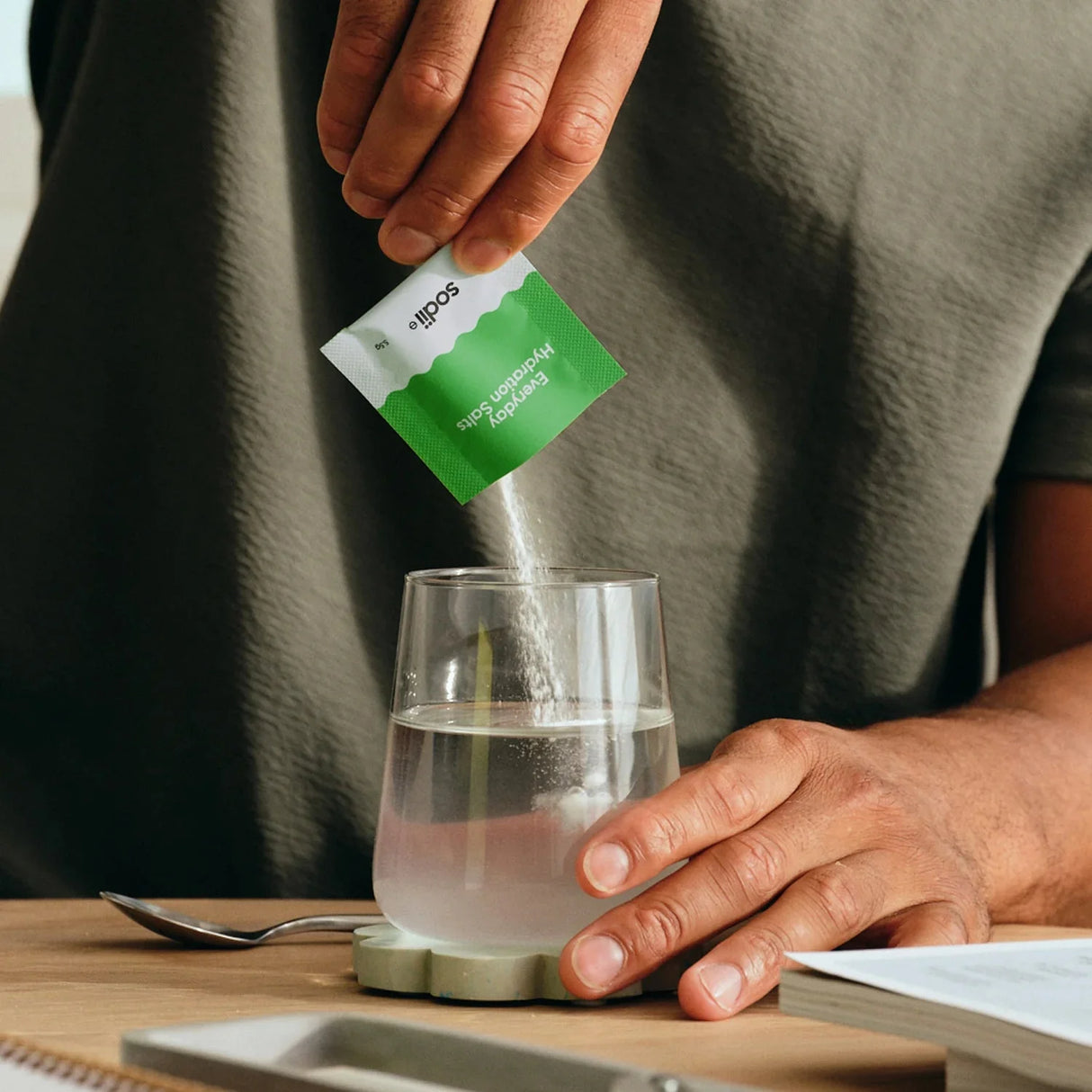 Hands pouring Salty Kiwi - 30 Sachets by Sodii electrolyte powder into glass water, on wooden table with book nearby