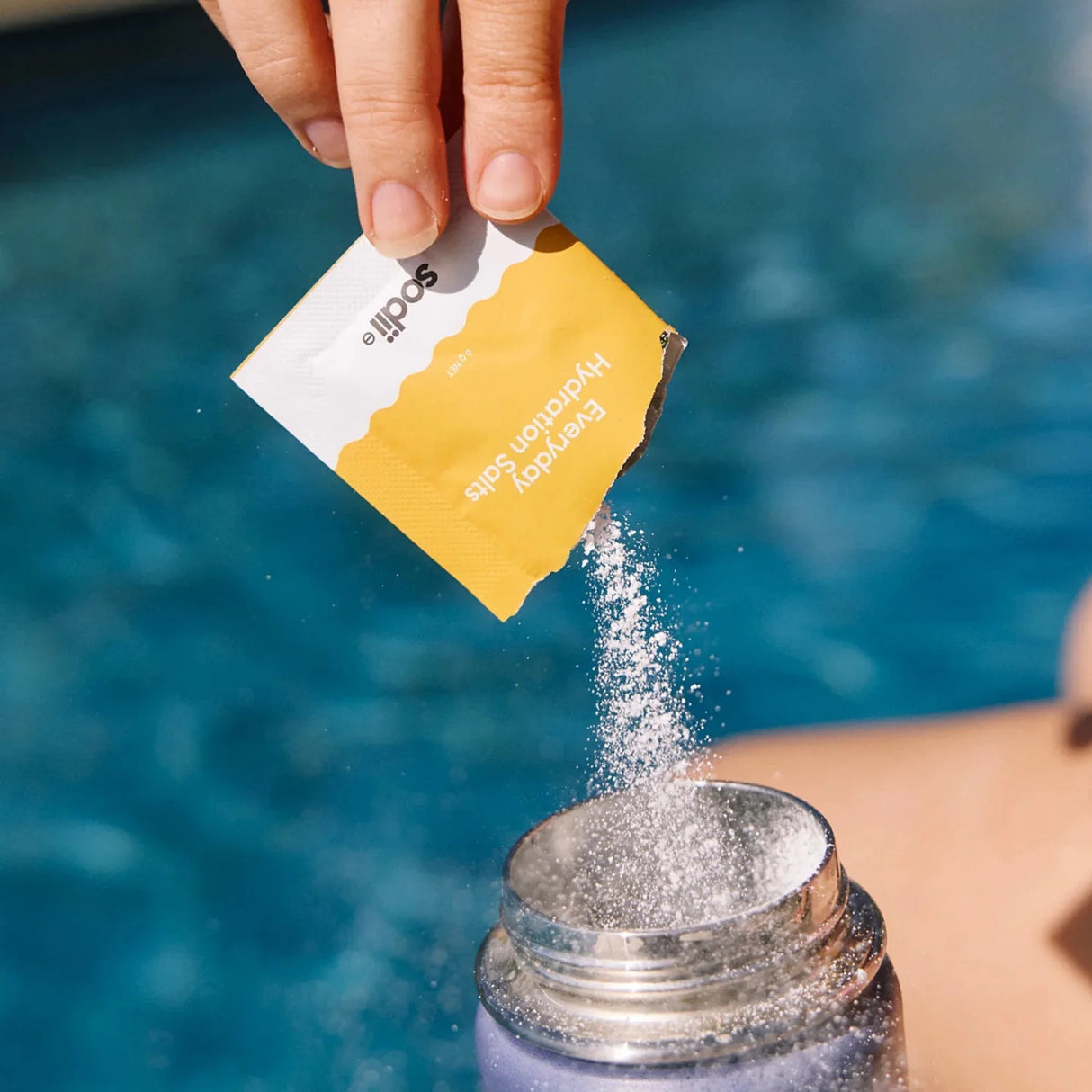 Hand pouring Salty Pineapple - 30 Sachets powder by Sodii into water bottle near pool backdrop