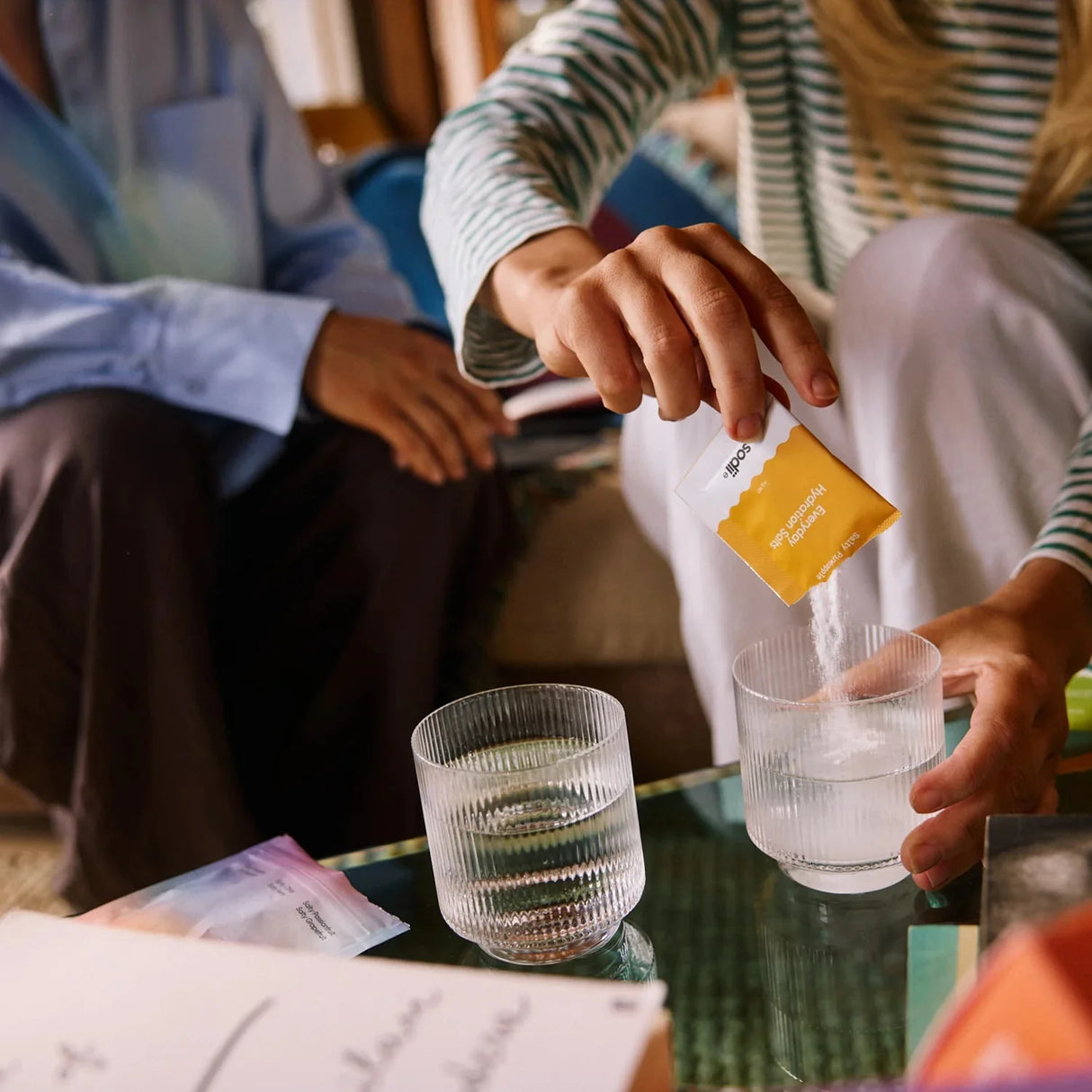 Person pouring Salty Pineapple - 30 Sachets by Sodii into glass of water, with another glass nearby on table.