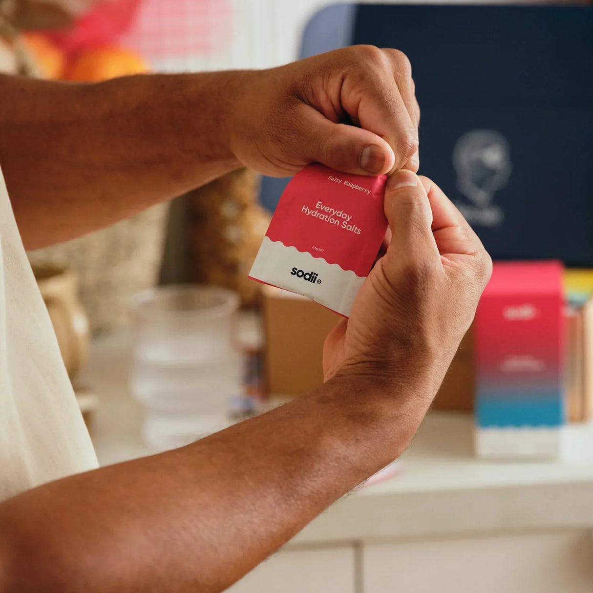 Person holding Salty Raspberry - 30 Sachets hydration salts by Sodii with blurred kitchen background and water glass