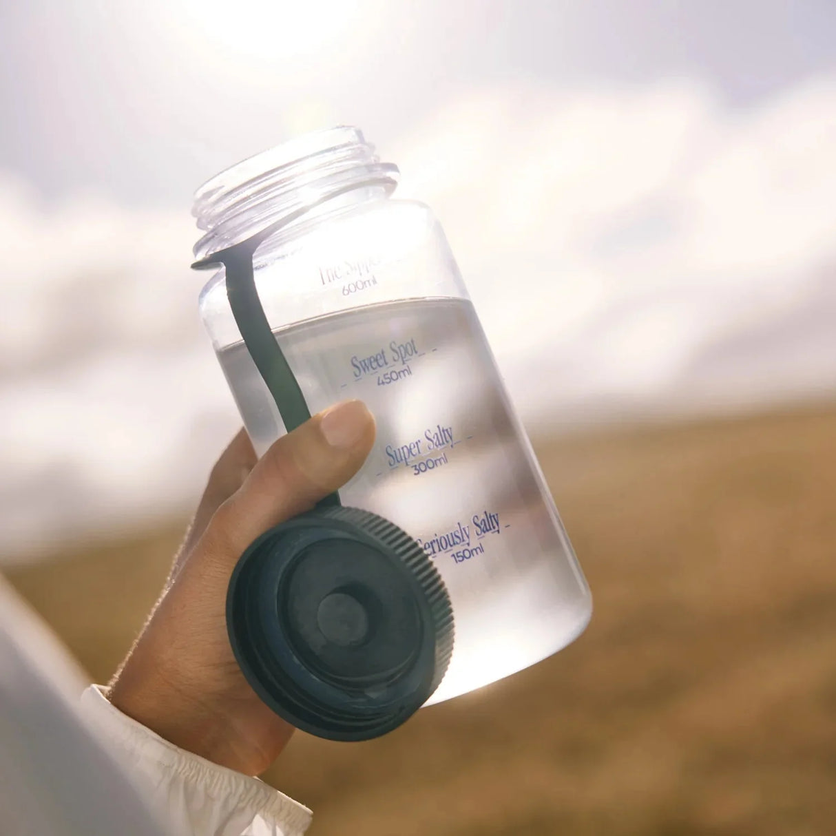 Hand holding clear water bottle with dark lid outdoors under cloudy sky, promoting Unflavoured - 30 Sachets by Sodii.