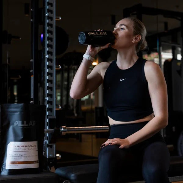 Woman in black workout clothes drinking from PILLAR PERFORMANCE shaker next to WPI - Chocolate - 1kg protein powder bag