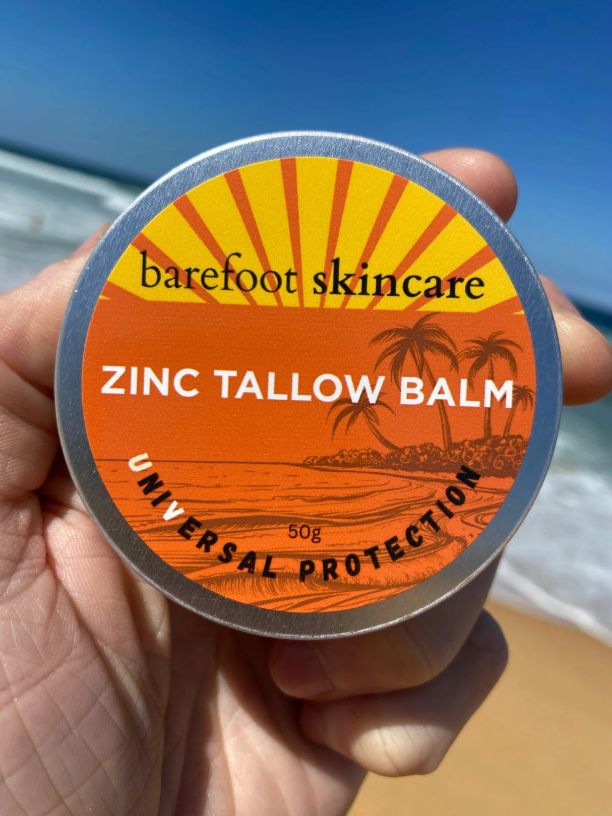 Hand holding Zinc Tallow Balm - 130g tin by Barefoot Skincare Co with beach and ocean waves in background
