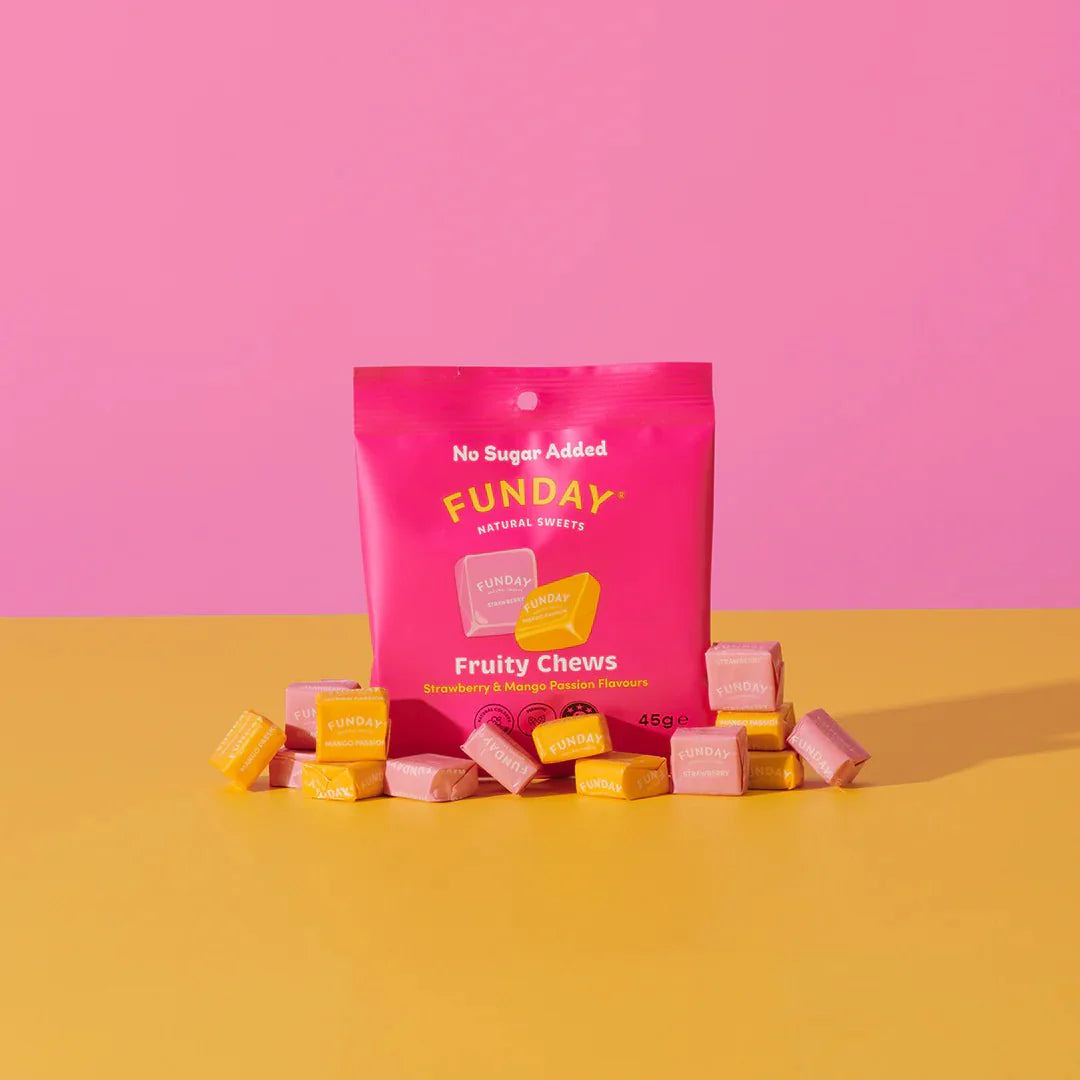 A bright pink Funday Fruity Chews - 45g packet, made with plant-based prebiotic fibre and no added sugar, sits on a yellow surface surrounded by individually wrapped yellow and pink candies, against a pink background.