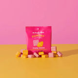 A bright pink Funday Fruity Chews - 45g packet, made with plant-based prebiotic fibre and no added sugar, sits on a yellow surface surrounded by individually wrapped yellow and pink candies, against a pink background.