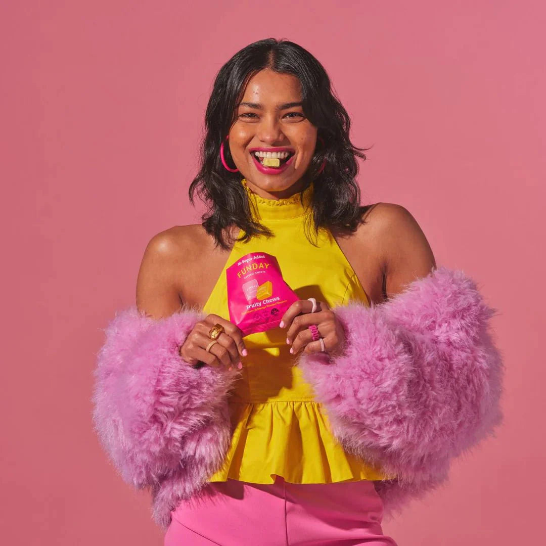 A smiling woman in a yellow top and pink attire holds a Funday Fruity Chews - 45g bag with plant-based prebiotic fibre and no added sugar, standing against a pink background, accessorized with pink hoop earrings and rings.