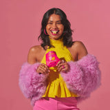 A smiling woman in a yellow top and pink attire holds a Funday Fruity Chews - 45g bag with plant-based prebiotic fibre and no added sugar, standing against a pink background, accessorized with pink hoop earrings and rings.