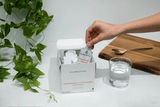 Hand holding a pouch of CYMBIOTIKA Magnesium L-Threonate - 30 Pouches supplement next to a glass of water and green plant.