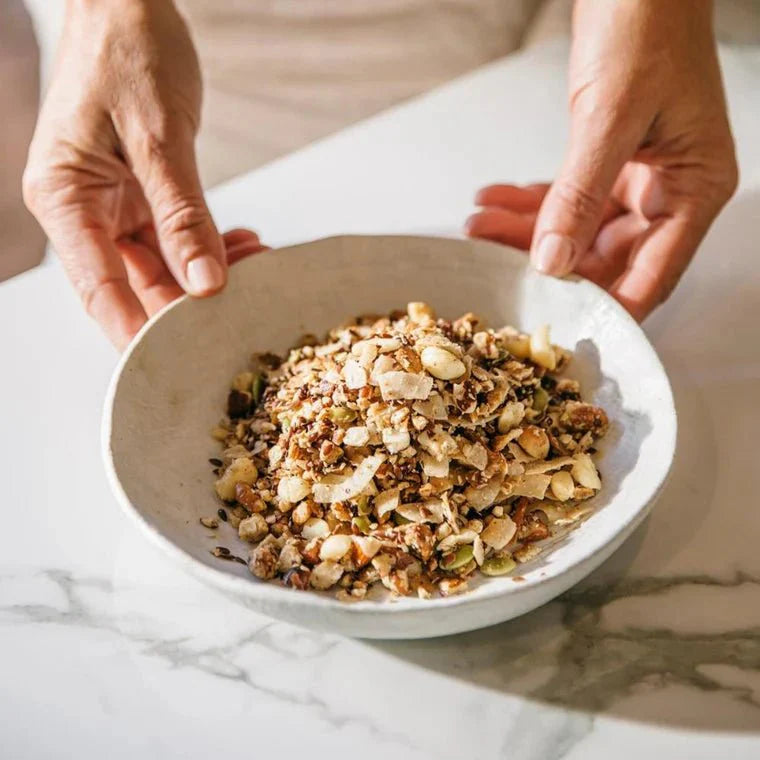 Hands holding a bowl of Keto Premium Paleo Granola - Macadamia & Coconut - 300g by Brookfarm on marble surface