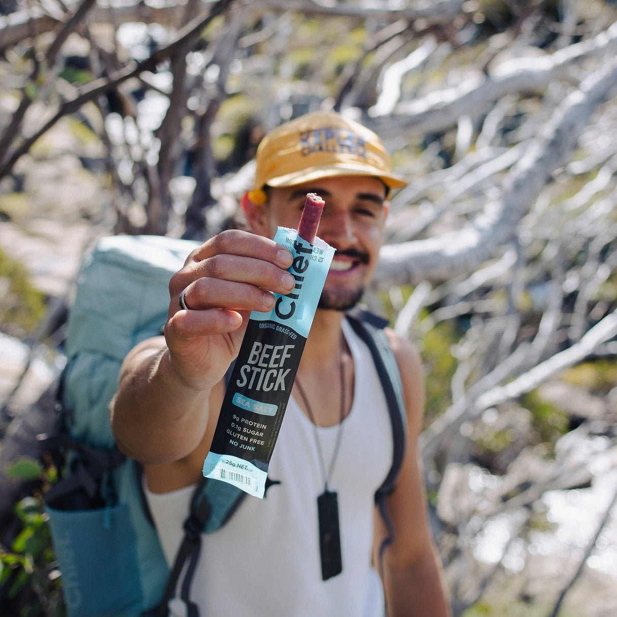 Man outdoors holding a Chief Organic Beef Stick - Sea Salt - 25g with backpack and trees in background