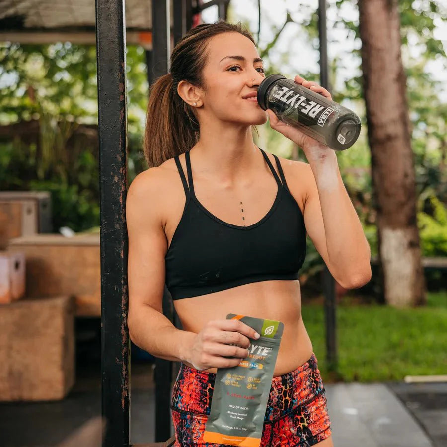 Woman in workout gear drinking from Re-Lyte bottle and holding Re-Lyte Pre-Workout Sample Pack - 4 Sticks outside gym area