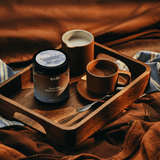 Wooden tray holding a sodii Salty Chocolate - 30 Serve Tub next to a cup of coffee and a cream pitcher, set on a cozy brown fabric background.